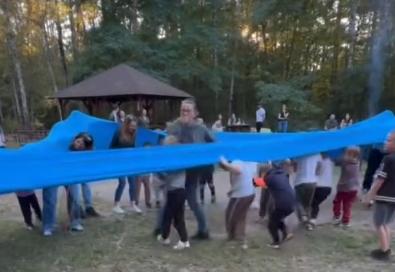 Dzieci w grupie otoczone są niebiską gumą i próbują ją rozciągnąć.
