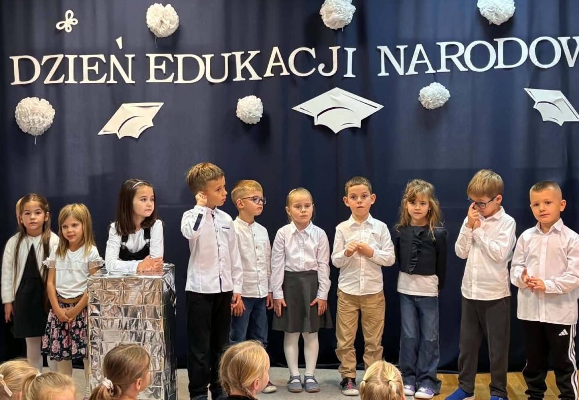 Dzieci ubrane na galowo stoją w półkolu na tle napisu dzień edukacji narodowej i recytują wierszyki.