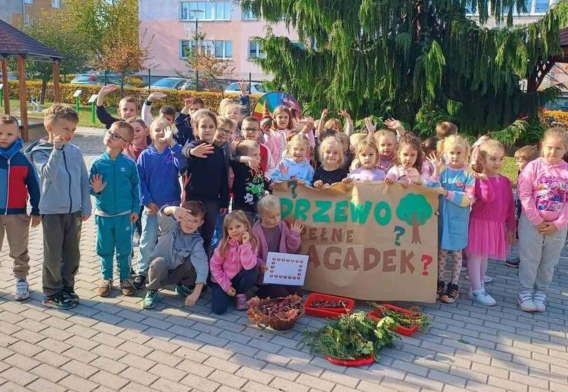 Dzieci stoja w grupie na dworze trzymając w ręku plakat z napisem drzewo pełne zagadek.