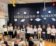 Dzieci ubrane na galowo stoją w półkolu na tle napisu dzień edukacji narodowej i recytują wierszyki.